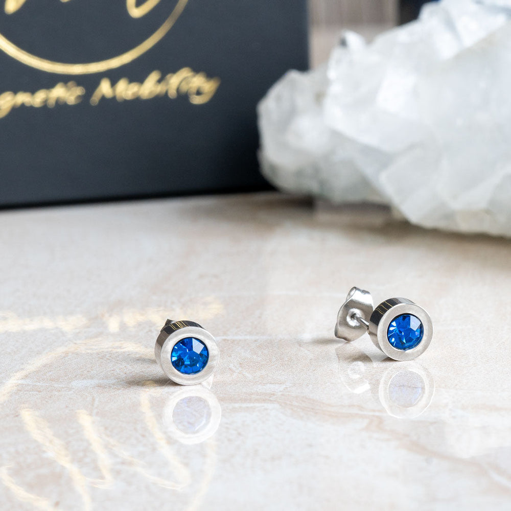 
                  
                    Load image into Gallery viewer, Birthstone Stud Earrings by Magnetic Mobility with blue Swarovski crystals. These earrings have a magnetic back and are ideal for fashionable pain relief, displayed on a reflective surface with Magnetic Mobility packaging.
                  
                