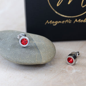 
                  
                    Load image into Gallery viewer, Magnetic Mobility Birthstone Stud Earrings featuring red Swarovski crystals. These earrings have a magnetic back and are ideal for providing stylish pain relief. Displayed on a smooth stone with Magnetic Mobility branded box in the background.
                  
                