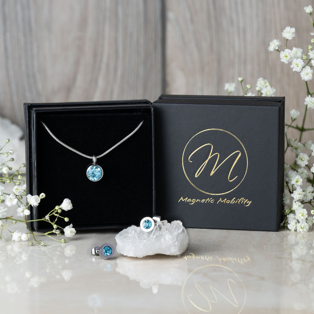 
                  
                    Load image into Gallery viewer, Magnetic Mobility March Birthstone gift set featuring a light blue aquamarine Swarovski crystal necklace and matching stud earrings. Each piece includes a magnetic back for stylish pain relief, beautifully presented with white flowers and a black branded box.
                  
                